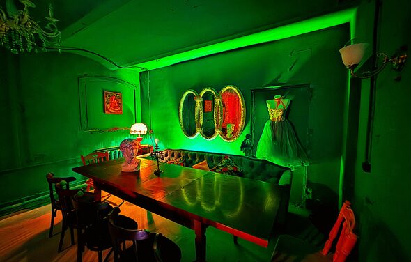 Лофт «Рич» (RICH). Зеленый интерьер с длинным деревянным столом, окруженным черными стульями. На столе лампа с абажуром. На стене три зеркала овальной формы. Угловая софа с подушками. Платье на манекене. Зеленая подсветка. Потолочный светильник.