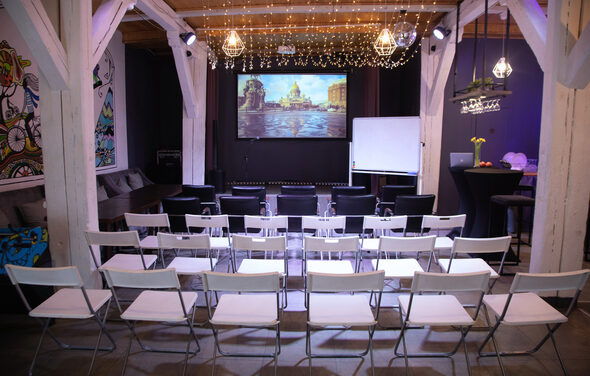 Сатурн Лофт. Многофункциональное помещение с деревянными балками, экраном и проектором. Зал оформлен стульями в несколько рядов, на стене вывешен белый экран. Имеются черные стулья, белая доска и высокий стол с цветами. Потолок украшен гирляндами и светильниками.
