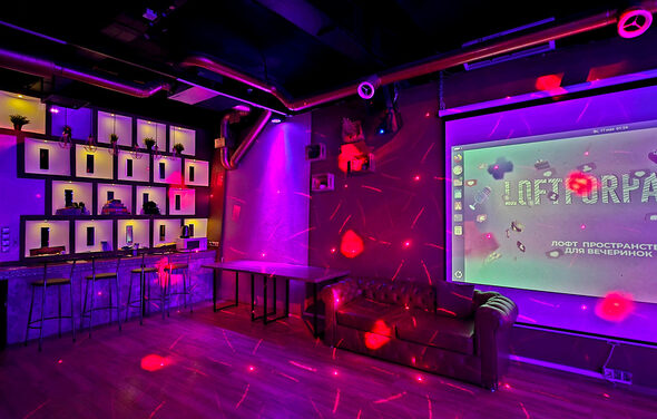 LoftForParty. Бэкстейдж лофт . Арендное пространство с низким баром и стульями, настроенной полкой с подсветкой. Рядом находится диван у стены с проекционным экраном. Освещение разноцветное, пол деревянный. Стены тёмные с декоративными элементами.