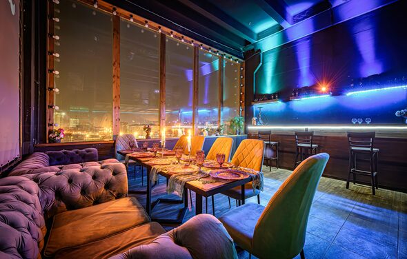 Лофт ZUM(Зум). Полузатемнённое помещение с панорамными окнами, обставленное мягкими диванами и стульями. Длинный стол покрыт дорожкой, на которой расставлены бокалы и тарелки. Яркое разноцветное неоновое освещение, стены сине-фиолетовые, пол серый.
