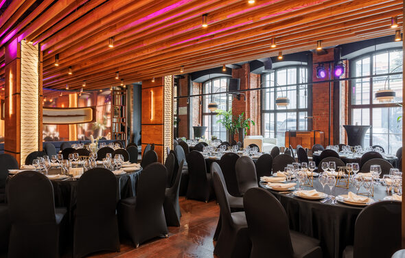 Zafferano Loft(Зафферано лофт). Банкетный зал с круглым столами, покрытыми черными скатертями и сервировкой из бокалов и тарелок. Кресла в черных чехлах. Потолок с деревянными балками и встроенным освещением. Стены из кирпича с большими арочными окнами.