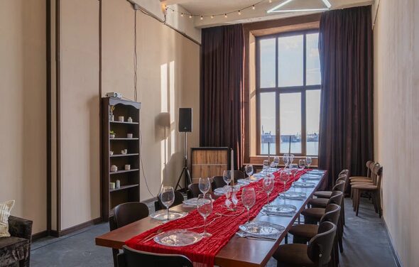 Chocolate Loft(Шоколад Лофт). Зал с видом на Финский залив. Просторное помещение с длинным столом и стульями. На столе красная дорожка, стеклянные бокалы и тарелки. Большое окно с тёмными занавесками. На стенах полки с посудой, акустическая колонка и проектор. Серые полы, светлые стены, подвесное освещение.