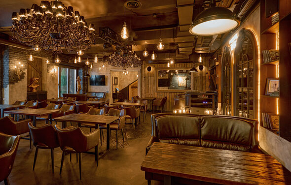 Tangiers Lounge(Танжер Лаундж) . Помещение с деревянными столами и кожаными креслами. Подвесные светильники и люстры создают тёплое освещение. Стены декорированы картинами и прямоугольным зеркалом. Пол из тёмного материала. На стене полка с книгами. Потолок темный, с проводкой и вентиляцией.