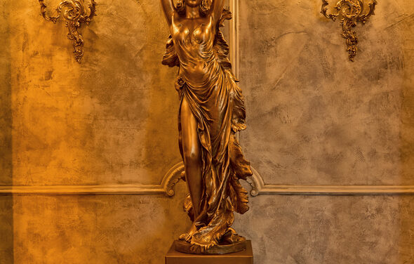 Tangiers Lounge(Танжер Лаундж) . Золотая скульптура девушки с поднятыми руками на фоне стены с декоративной штукатуркой серо-бежевого цвета, украшенной лепнинами. По бокам стены расположены две настенные лампы в классическом стиле с резными элементами. Освещение теплое.