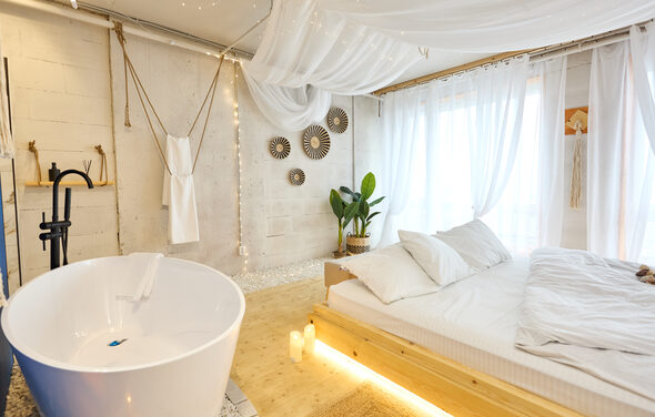 Пляжный лофт с галькой, открытой ванной, панорамными окнами. Просторная светлая зона с ванной и кроватью. Ванная белая, отдельно стоящая. Кровать с деревянным каркасом, белое постельное белье. Светло-бежевые стены с декоративными элементами. Растение в углу, на окнах белые шторы, потолочные тканевые занавеси.