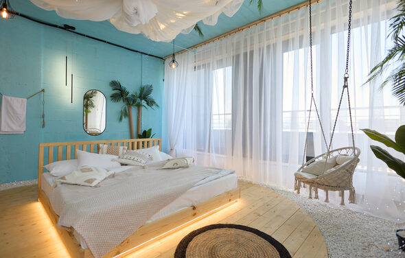 Лофт Мальдивы с открытым душем, балконом и видом на Сити. Спальня с деревянной кроватью на круглой платформе и подвесным креслом. Голубые стены, белые шторы, зеркало, небольшие растения. Натяжной светлый потолок с текстилем, светильники. Нейтральное постельное белье, коврик, белый и песочный цвета.