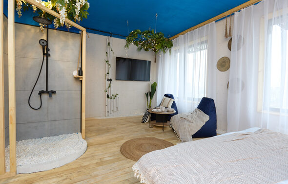 Лофт Бали с фото зоной на 23 этаже с видом на Москва-реку. Просторная комната с деревянным полом и синим потолком. Душевая зона с серыми плитками и черной сантехникой, рядом кровать с покрывалом, два кресла-мешка, телевизор на стене, прозрачные шторы. Декор из подвесных растений, коврик.