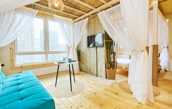 Тропический лофт с бунгало, баром и французским балконом. Жилая комната с деревянным полом и потолком, оформленным из бамбука. Синий диван, круглый столик с двумя стаканами. Светлые шторы разделяют зону с кроватью. Декор растениями в горшках. Мягкое дневное освещение из окна, телевизор на стене.