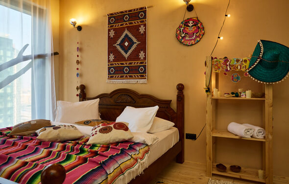 ⁠Лофт в Мексиканском стиле для костюмированных вечеринок. Спальня с деревянной кроватью, украшенная цветным покрывалом и подушками. Стены в бежевых тонах с декоративными элементами, полка с декоративными предметами и полотенцами. Освещение от настенных бра. Белые занавески на окне.