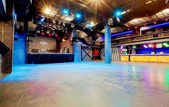 PRAVDA(Правда). Просторный зал на 800 гостей. Широкое внутреннее пространство с бетонным полом. Площадка с оборудованием для выступлений. Стойка бара с подсветкой. Освещение разноцветное, включая прожекторы и неон. Высокий потолок с открытыми металлическими конструкциями. Тёмные стены.