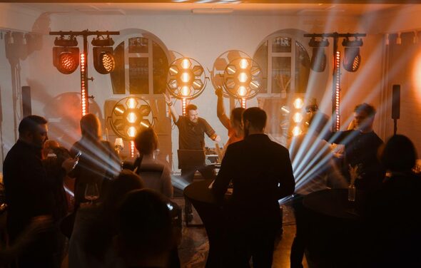 Светлый Лофт. Пространство для вечеринки с диджеем, круглыми высокими столами, людьми, световыми установками и эффектами. Белые кирпичные стены, большие окна, оранжевое освещение, черные мебельные чехлы.