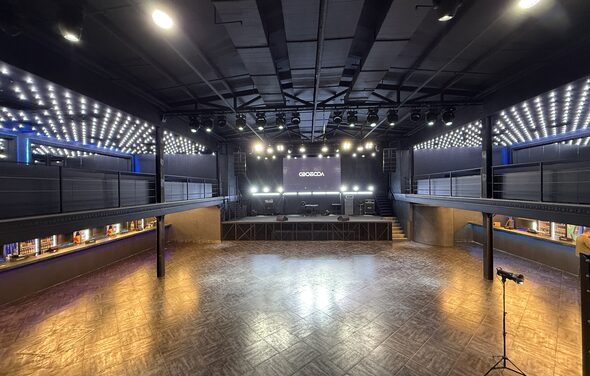 СВОБОДА. Зал на 1200 человек. Просторное темное помещение с паркетным полом, центральной сценой и балконами по обе стороны. Разнообразное освещение, включая потолочные светильники и светодиодные полосы. На сцене черная стена с надписью. Справа микрофон на стойке.