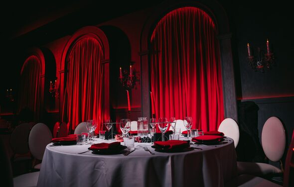 QUQLA. Круглый стол с белой скатертью, сервирован черной и красной посудой; бокалы и столовые приборы. Белые стулья с округлыми спинками. Красные шторы, подсвеченные снизу. Темные стены с арочными нишами и канделябрами.