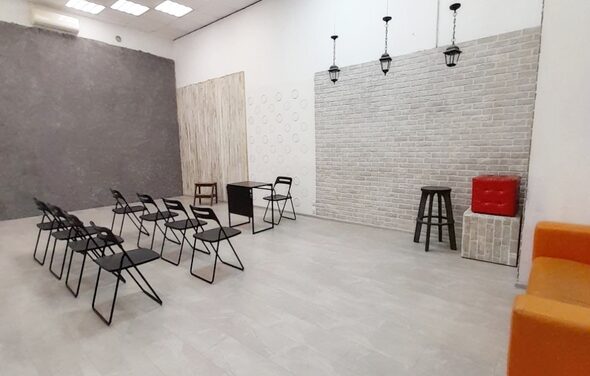 Интер-Фото. Просторное помещение с кирпичной и бетонной стенами, расположенными под углом. На полу светлая плитка. В центре черные складные стулья в два ряда с небольшим столом. Подвесные светильники. По краям стула, стол, пуф и барный стул.
