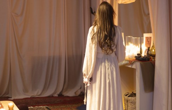 Лофт Поле. Уютный зал для мероприятий. Текстильное пространство с навесными тканями, журнальный столик с зажженными свечами и декором, штативная лампа, мягкие ковры с восточными узорами, светлая одежда человека, мягкое освещение.