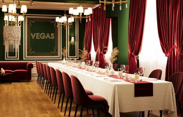 Вегас. Атмосферный лофт с акцентным дизайном. Банкетный зал с длинным столом, накрытым белой скатертью и красными салфетками, окружённый бордовыми стульями. Красные бархатные шторы и зелёные стены. Светильники с подсвечниками. На стене текст: "VEGAS". Пол из светлого дерева.