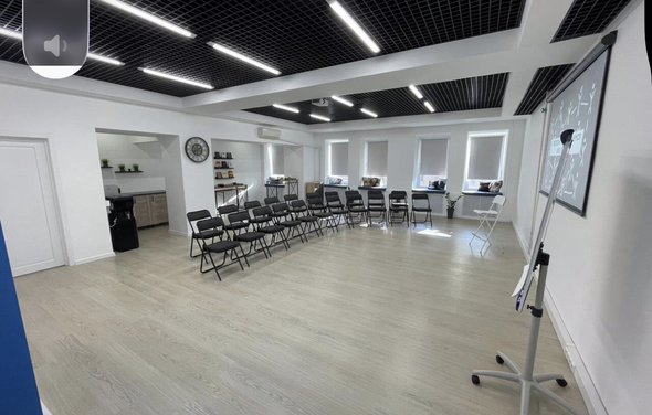 Уютный тренинговый зал на Таганской. Просторное помещение в светлых тонах, с деревянным полом, белыми стенами и потолками с черными решетками и встроенным освещением. Ряды складных стульев, магнитно-маркерная доска на стенде, кухня с посудой слева, окна с жалюзи.