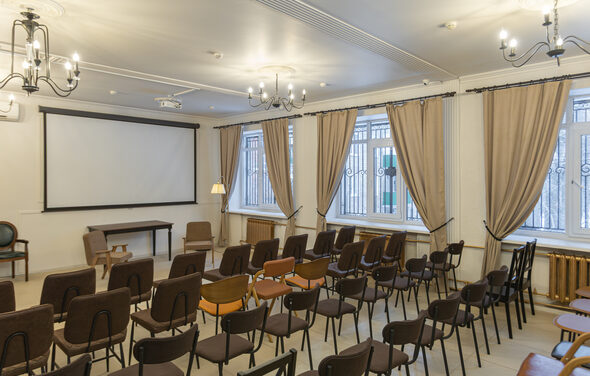 Open Space (Открытое пространство) для мероприятий. Конференц-зал с рядами стульев, деревянными полами и белыми стенами. Экран для проекции, столы и кресла. Светильники на потолке и торшер. Большие окна с бежевыми шторами. Металлические решетки на окнах, светлое освещение.