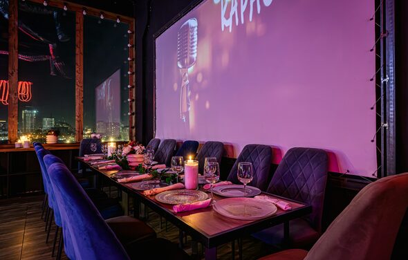 Лофт Овертон. Уютное помещение с длинным деревянным столом и бархатными стульями. Настольные украшения, свечи и посуда на столе. На стене большой экран или панель с изображением микрофона. Светодиодные гирлянды и окна с ночным видом на город.