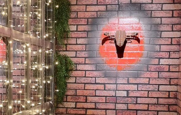 BULL LOFT(Лофт Бык) . Кирпичная стена с граффити в форме красного круга и черной гитары. Слева деревянная конструкция с висящими гирляндами и зеленью. Освещение теплое, мягкое.