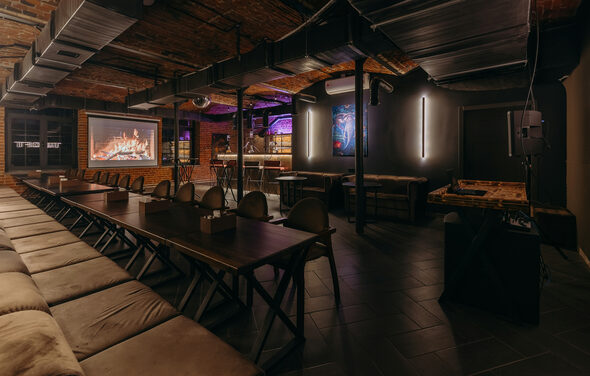 Авторские пространство T&S loft (ти энд си лофт) . Просторное помещение с кирпичными стенами и деревянным потолком, длинные деревянные столы и скамейки, игровые столы, проектор с изображением камина, настенные светильники, тёмные плиточные полы, зона с барной стойкой и неоновой подсветкой.