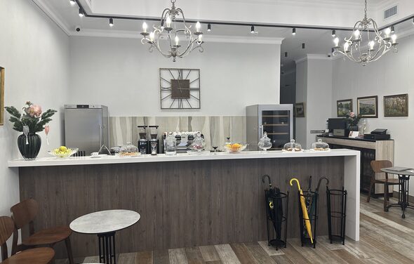 Арт Холл. Кафе с деревянной барной стойкой, круглые столы и стулья, светильники-подвески, светлые стены, картины, металлические зонтовницы с зонтами, кофемашина и напитки на стойке, декор в виде часов, деревянный пол.