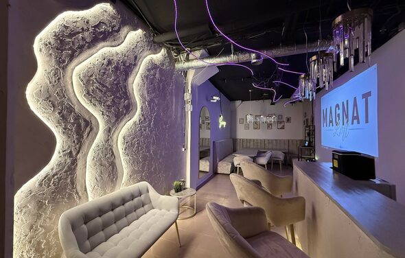 Лофт SUNRISE(Восход Солнца). Просторное помещение на 15 гостей. Лофт пространство с неоновым и белым освещением, выделяющим текстурированные стены. Мягкие стулья и диваны, столики. Барная стойка слева. Потолок с открытыми коммуникациями. На стене телевизор с надписью. Светильники свисают с потолка.