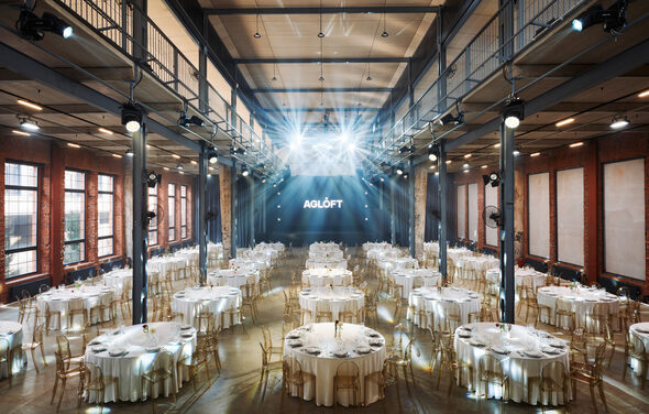AG LOFT. Grand Hall (АГ Лофт. Большой зал) . Большое индустриальное пространство с высокими потолками и множеством круглых столов, покрытых белыми скатертями. Стулья прозрачные, на столах находятся свечи и бокалы. Огоньки сверху и кирпичные стены. На стене текст: "AGLOFT".