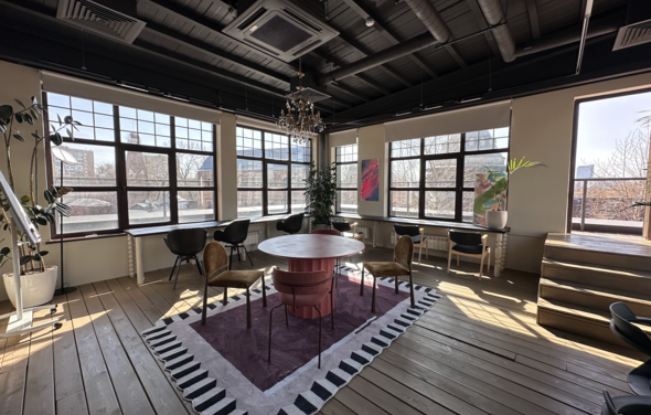 Пространство с террасой до 50 человек. Просторное помещение с большими окнами и деревянным полом. Центральный круглый стол окружен стульями на ковре с геометрическим узором. Подвесная люстра на черном потолке с видимыми трубами. Растения и картины по стенам.