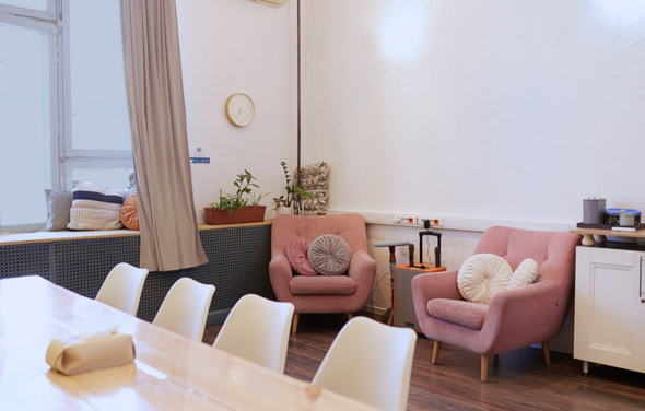 H4U. Светлый зал П-602. Современное пространство с панорамными окнами и кремовыми шторами. Обеденный стол из светлого дерева с белыми стульями. Два розовых кресла с подушками. Декор из растений и скульптур. Белые стены, лампа на стене, настенные часы.