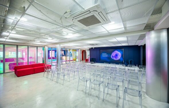 АКИ.лаб пространство для мероприятий. Современное концертное или презентационное пространство с прозрачными стульями, красными диванами, большим экраном на темной стене, бетонный пол, подвесной потолок с вентиляцией, разноцветная подсветка, стеклянные перегородки.