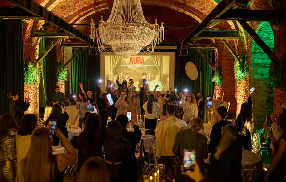 Arsenal Loft(Арсенал Лофт). Банкетный зал с кирпичными стенами и арочными потолками. Большая люстра, зеленые и оранжевые подсветки. Люди стоят у сцены. Круглые столы с белыми скатертями. На заднем плане экран с надписью "AURA".
