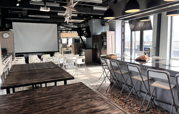 Зал под кулинарные мастер классы и мероприятия. Просторное помещение с деревянными столами и белыми стульями, большой экран на передней стене. Справа барная стойка с высокими стульями. Потолок с подвесными светильниками. На полу плитка с узором. Есть большие окна.