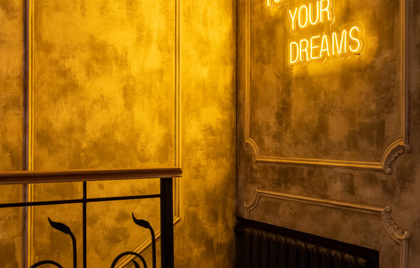 Каминный Зал Tangiers lounge Pokrovka(Танжерс Лаундж Покровка). Зона с декоративным металлическим ограждением и неоновой надписью на стене, текст читается частично. Стены с фактурной отделкой серого цвета, освещение теплое, желтоватое. Черный радиатор под стеной.