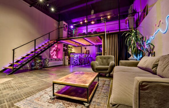 НЕОН Лофт. Лофт с антресолью, стеклянные ограждения, лестница с металлическими перилами. Диван, кресло, низкий стол. Неоновая вывеска, растения. Фиолетовая и теплая подсветка. Кирпичные стены, кафельный пол. Барная стойка с граффити. Чёрные шторы.