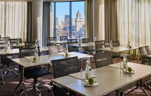 Конференц-зал Экспресс Махараджей. Конференц-зал с большими окнами и видом на город, расставлены прямоугольные столы с черными стульями на колесиках. На столах размещены бутылки воды, стаканы и зеленые яблоки. Светлые шторы, ковровое покрытие.