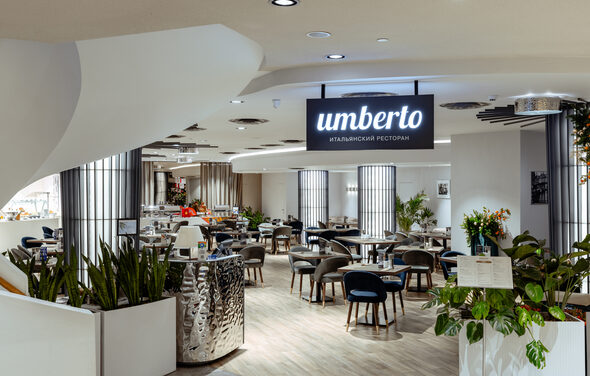 Итальянский ресторан Umberto(Умберто). Современное ресторанное пространство с круглыми столиками и мягкими стульями. Светлые деревянные полы, декоративные растения, светильники на потолке. На вывеске надпись «италиянский ресторан». Стойка из металла.