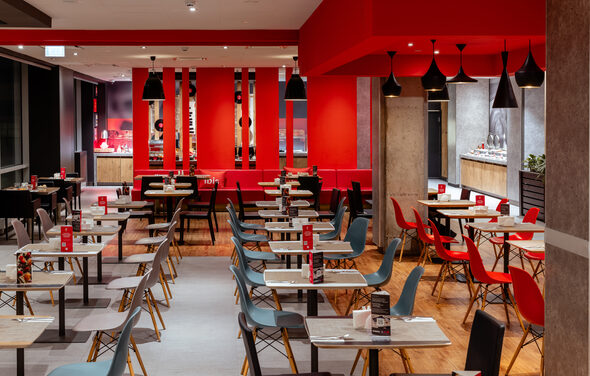 Ресторан Ibis Kitchen(Ибис Кухня). Современное кафе с открытой планировкой. Столы и стулья в нескольких рядах. Комбинация черных и серых стульев, столешницы светлого дерева. Акцентные красные детали, включая стены и стулья. Черные подвесные светильники. Пол из светлого дерева.