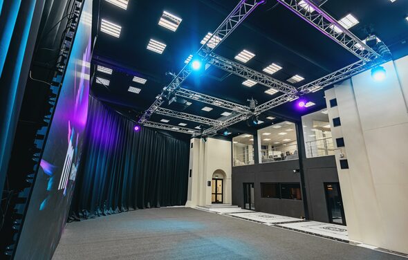 New Media (Нью Медиа) продакшн. Павильон с большим LED-экраном. Просторная комната с высокими потолками и черными занавесами. Освещение из встроенных потолочных светильников и прожекторов. Металлические конструкции для оборудования. Стены светлые, полы серые. Видны стеклянные перегородки и двери.