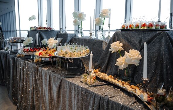 Арт-Плейс Фактура. Современный лофт с видом на весь город. Фуршетный стол с темной блестящей скатертью, сервированный закусками и десертами. Использованы возвышения для блюд. Декор из белых роз и свечей в подсвечниках. Большие окна на заднем плане обеспечивают естественное освещение.