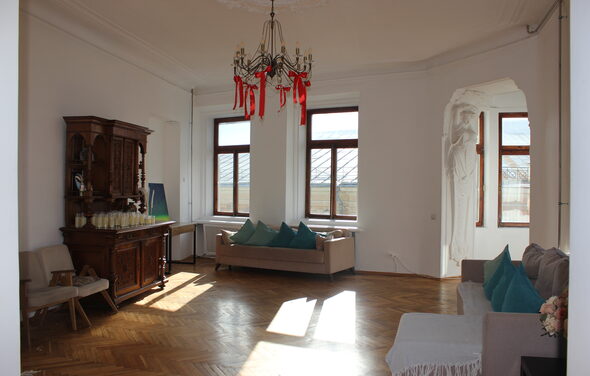 Cassiopea(Кассиопея). Просторная светлая комната с деревянным паркетом, белыми стенами и высокими потолками. Установлены два дивана и кресла с подушками. В центре — люстра с декоративными элементами. Несколько больших окон. Комод с напитками.