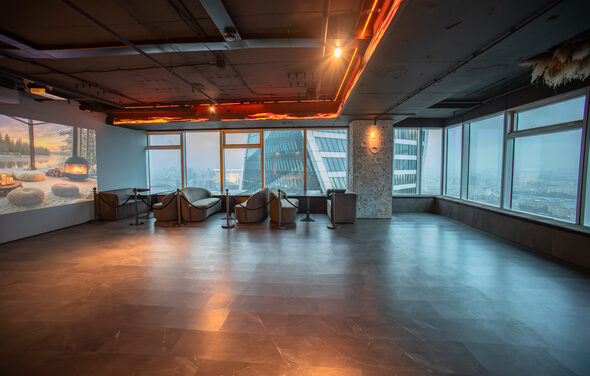 ATMOSFERA 53(Атмосфера 53). Просторное помещение с большими окнами, оклеенными изнутри жалюзи, вид на городские здания. Полы из темного материала, потолок с трассированными светильниками. Несколько стульев и столики у панорамных окон. На стене экран с изображением.