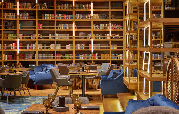Communitymoscow (Коммьюнити Москоу).Зал Библиотека. Многофкнкциональное пространство. Библиотечное пространство с высокими деревянными полками, заполненными книгами. Мягкие кресла серого и синего цвета вокруг круглых столов. Теплое освещение с вертикальными световыми полосами. На столах стеклянные бокалы янтарного цвета.