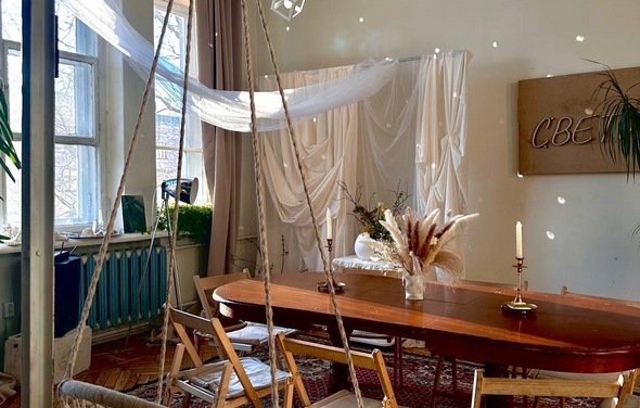 Студия Свети. Просторная комната с деревянным столом и складными стульями. На столе свечи и композиция из сухих трав. Стена декорирована светлой тканью, на стенде надпись кириллицей. Окна с прозрачными и светлыми шторами, зелёные радиаторы.