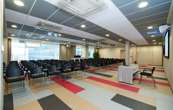 Конференц-зал ТехноПрогресс. Конференц-зал с рядами черных стульев, проекторы на потолке, окно с плотными шторами, стол с белой скатертью, разноцветное ковровое покрытие в серых и красных тонах, светло-серые стены, встроенные светильники.