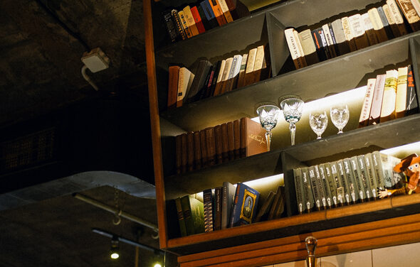 Гастробар Дневник. Книжная полка с разноцветными книгами и бокалами для вина, освещенная встроенной подсветкой. Потолок из темного бетона, видны элементы освещения и вентиляции. Доминируют теплые тона.