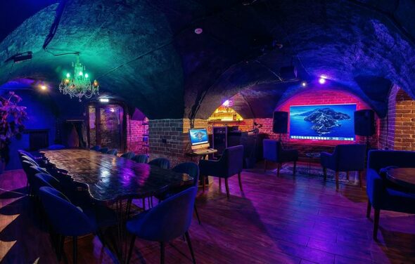 GB Rooms(ГБ Румс). Лофт №1. Подвальное помещение с арочными кирпичными стенами и потолками. Длинный деревянный стол с синими стульями. На стене большой экран, по бокам колонки. Декоративная люстра. Пол деревянный, освещение разноцветное. Компьютерный монитор на столе.
