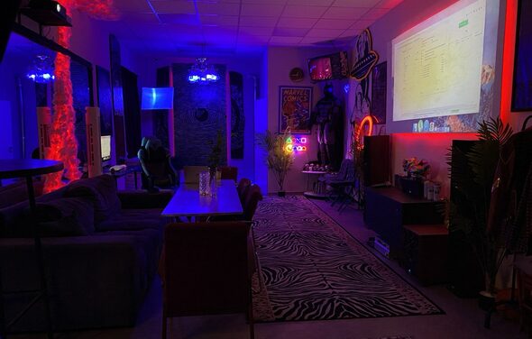 GB Rooms(ГБ Румс). Лофт №2. Комната с диванами и креслами, прямоугольный стол, проектор с экраном. Яркое разноцветное освещение, неоновые вывески, настенные постеры. Ковёр с зебровым узором, растения в горшках. Потолочные светильники, мебель из тёмных материалов.