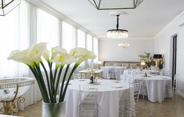 Лофт Космос. Зал Президент. Светлое банкетное пространство с белыми стенами и большими окнами, украшенными шторами. Круглые столы с белыми скатертями и прозрачными стульями. Потолочные люстры и настольные лампы создают освещение. В центре композиции белые каллы в вазе.