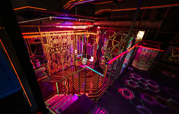 Лофт Космос. Зал Ночной Клуб. Креативное пространство с ярким неоновым освещением и геометрическими узорами. Металлические конструкции и лестница вниз, разноцветные световые линии на стенах и потолке, стеклянные панели. Тёмный фон, акцент на фиолетовый и оранжевый цвета.